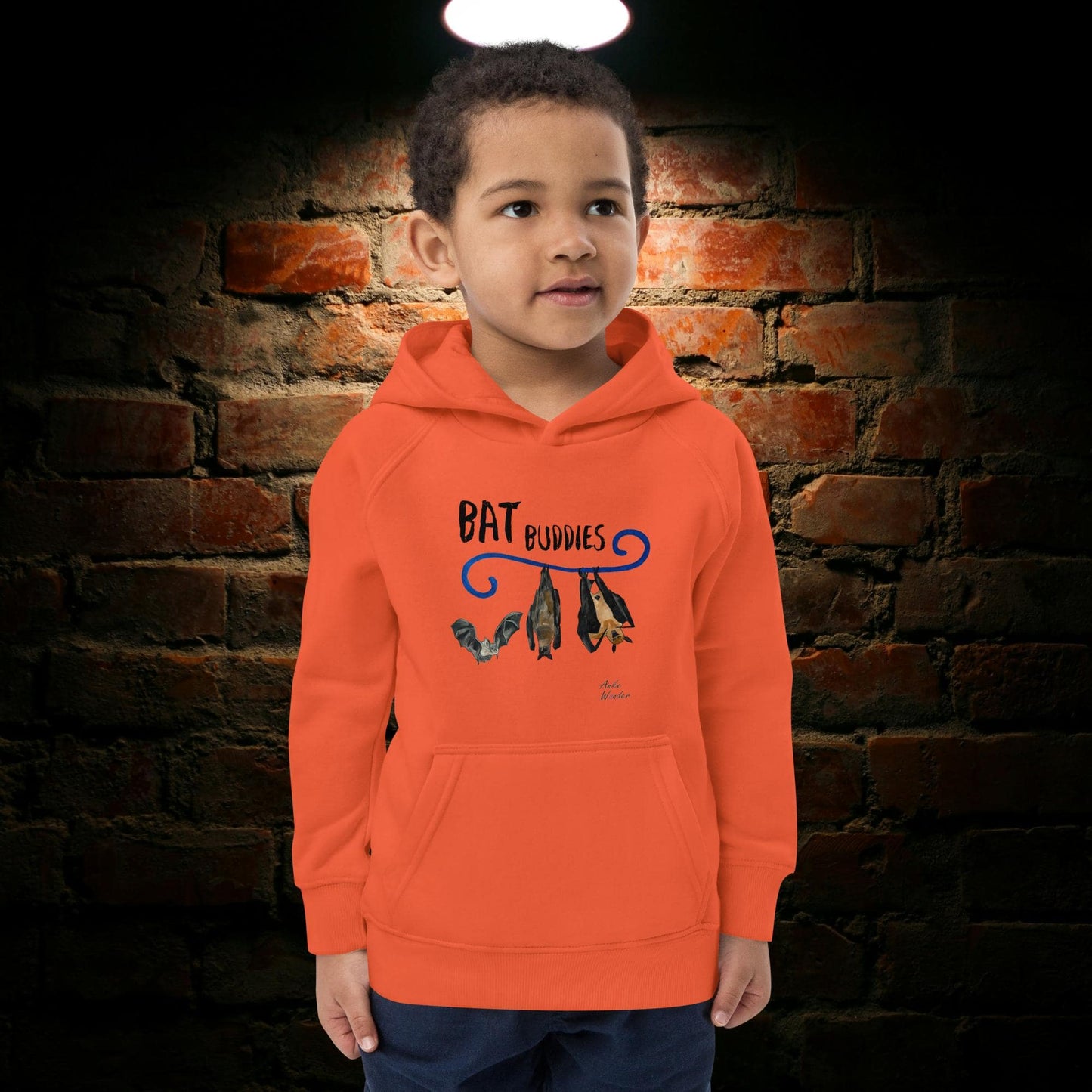 Boy wearing an orange hoodie with 'Bat Buddies' design against a brick wall.