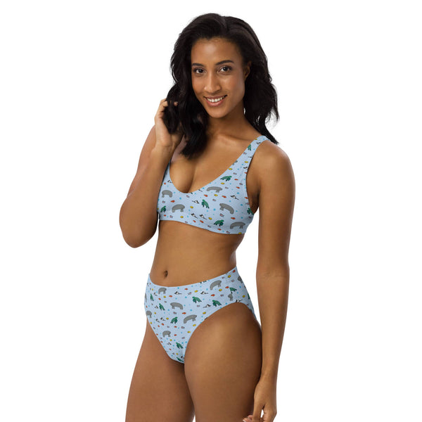 A woman models a high-waisted, eco-friendly bikini with a manatee and turtle print, captured from the front left angle to showcase the fit and design.