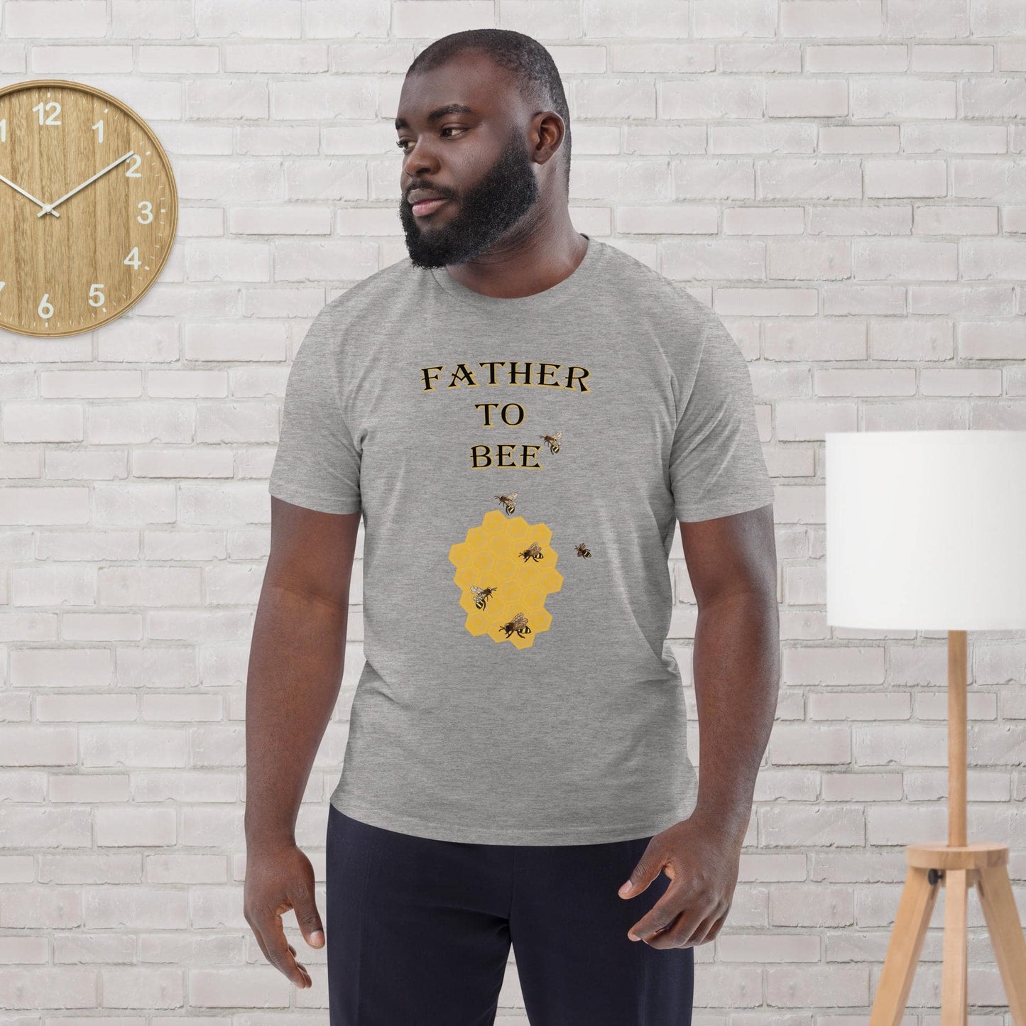 Man wearing a heather grey organic cotton t-shirt with “Father to Bee” text and bee graphic, standing in a modern room.