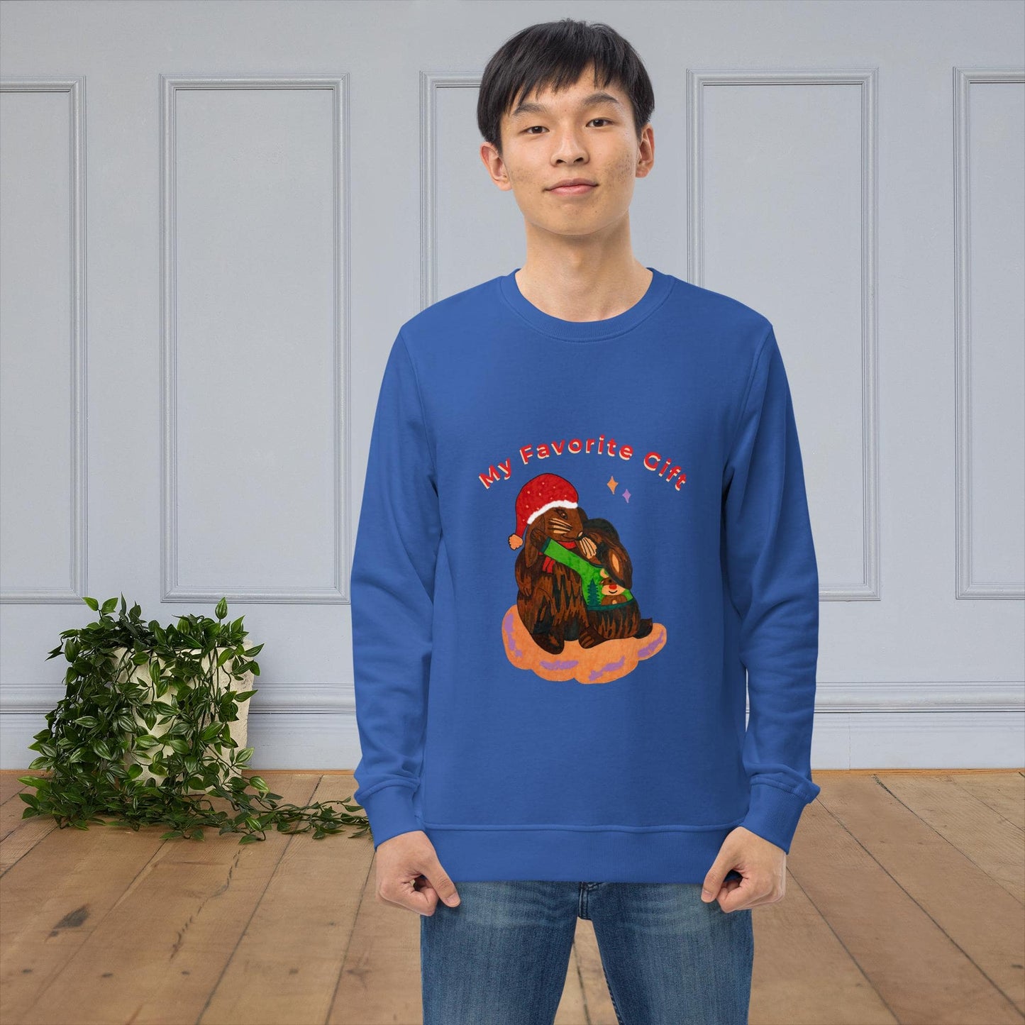 Man wearing a blue sweatshirt with a Christmas-themed bunny graphic and text 'My favorite gift' in a room with a plant and wooden floor.
