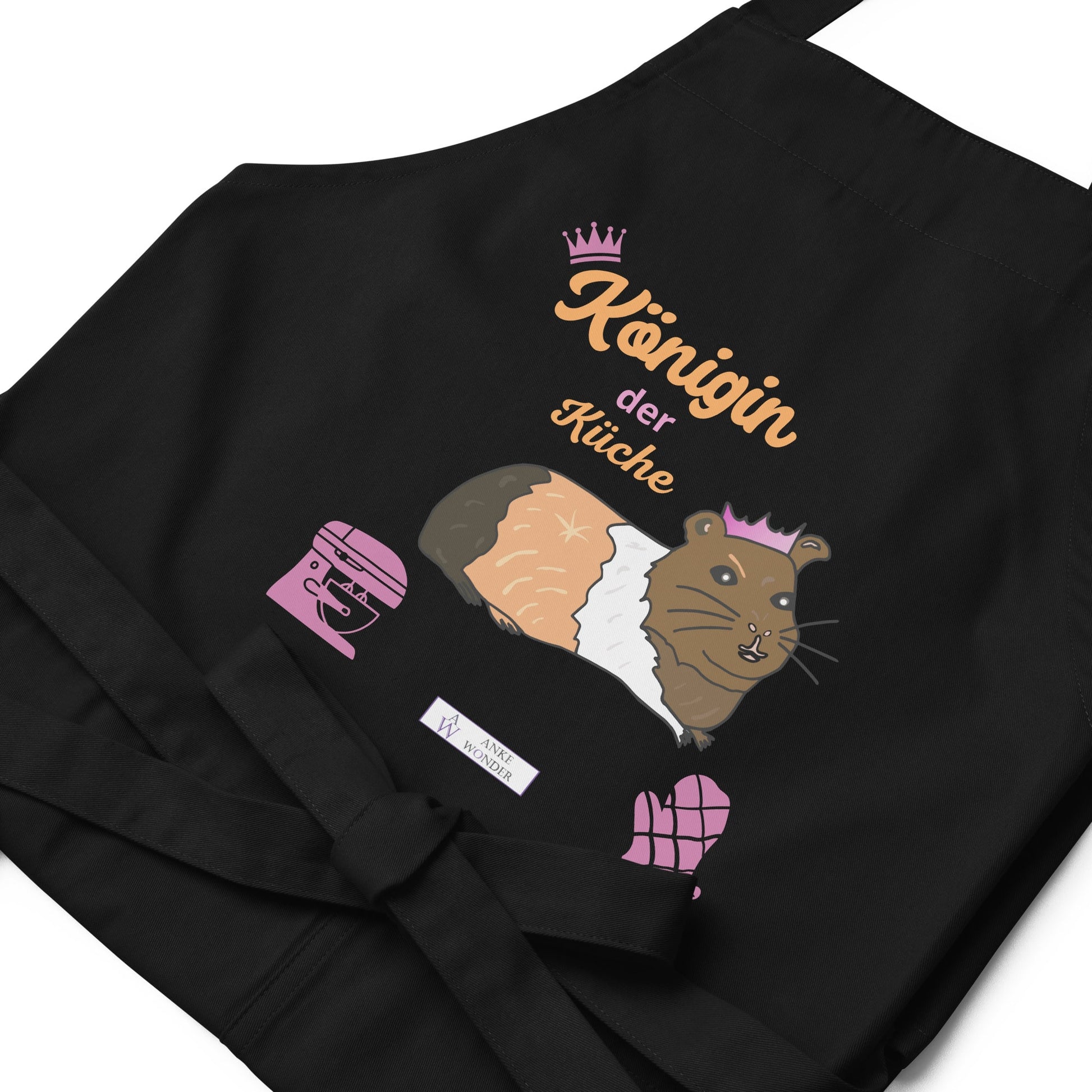 A flat-lay image of the black apron highlighting the detailed guinea pig illustration and the “Königin der Küche” lettering as well as the Anke Wonder branding.