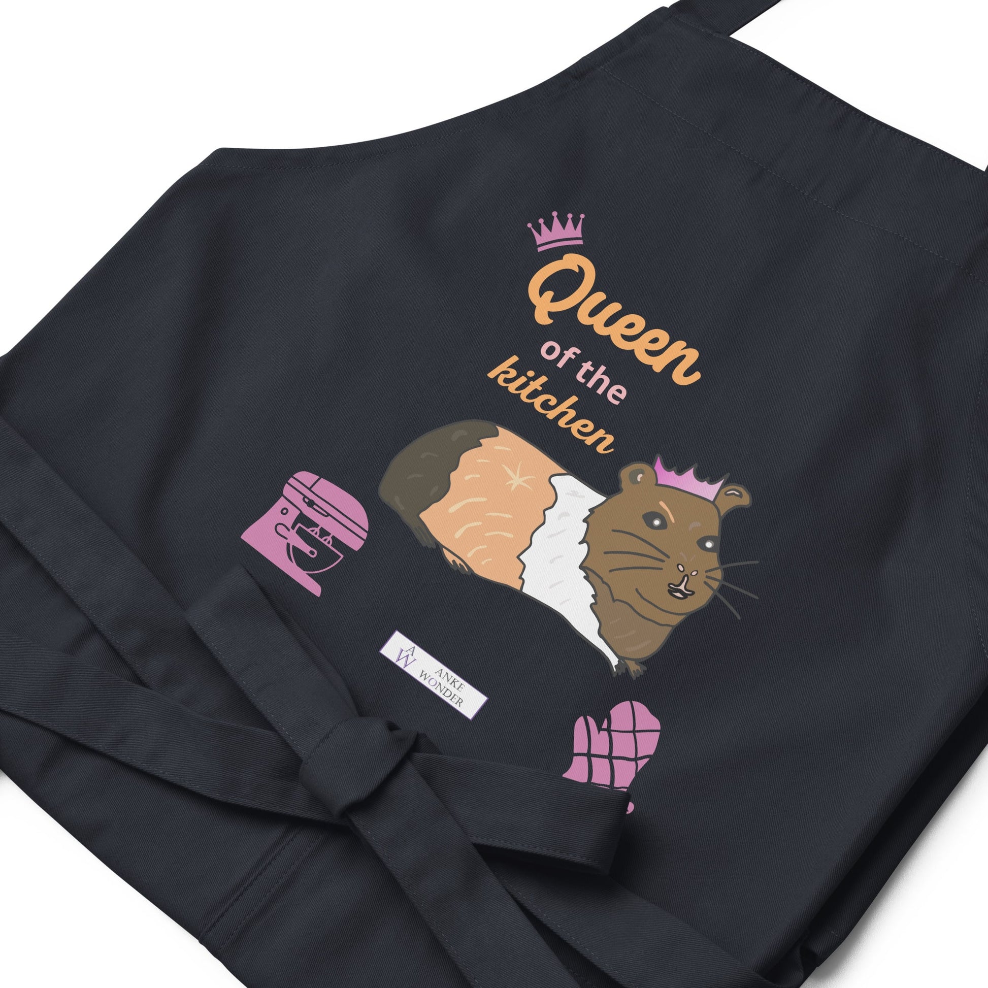 A flat-lay of the navy apron displaying the guinea pig in a pink crown and bold “Queen of the Kitchen” text with view of the Anke Wonder branding.