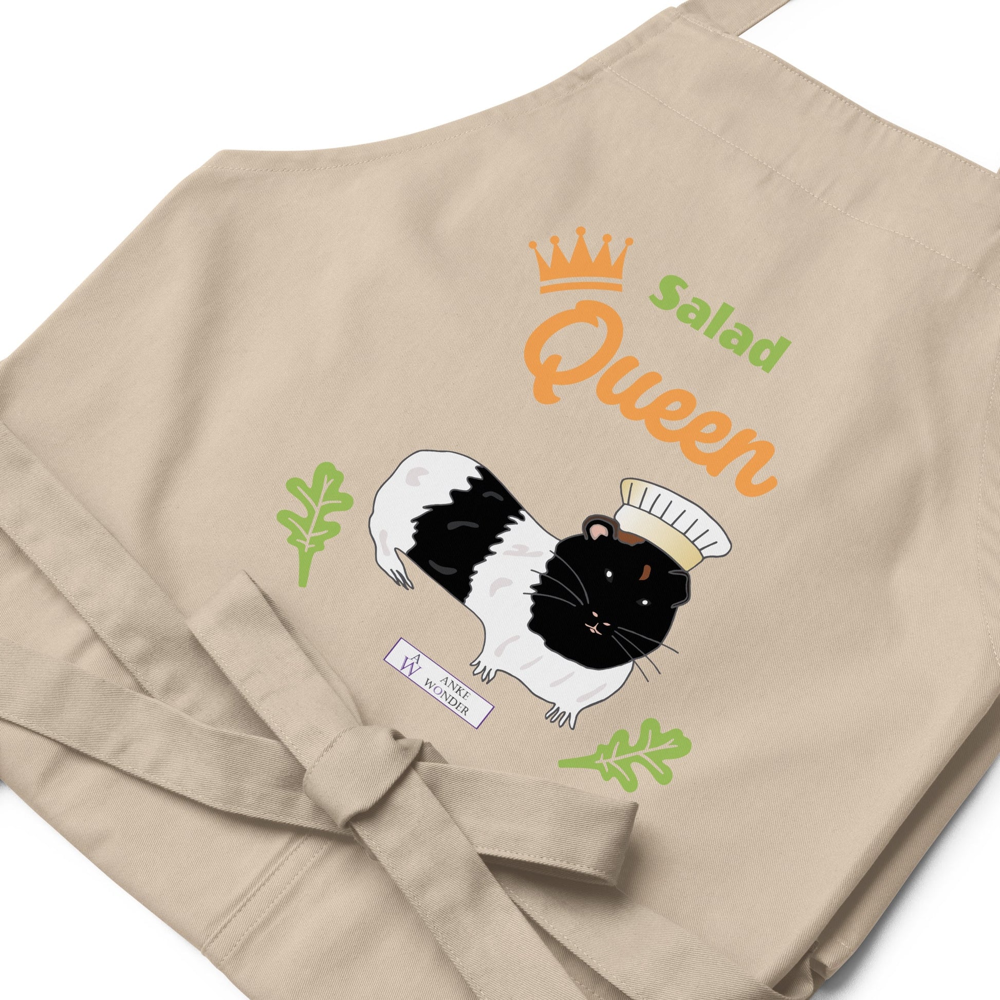 A flat-lay of the beige Salad Queen apron, showing the detailed guinea pig chef illustration and the Anke Wonder logo.