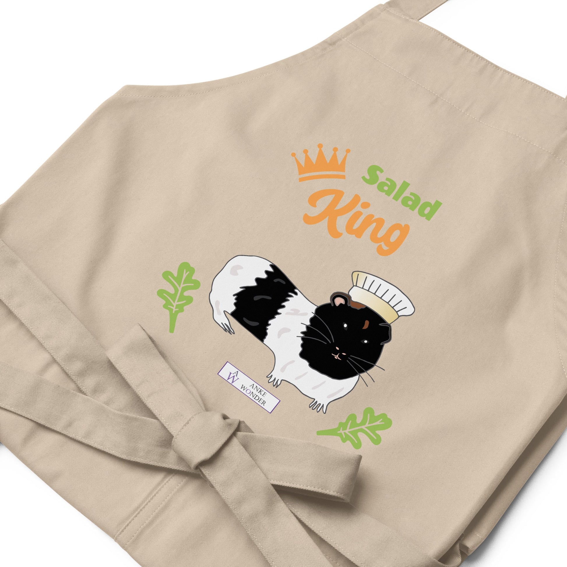 A flat-lay of the beige Salad King apron, showing the detailed guinea pig illustration and the Anke Wonder Logo.