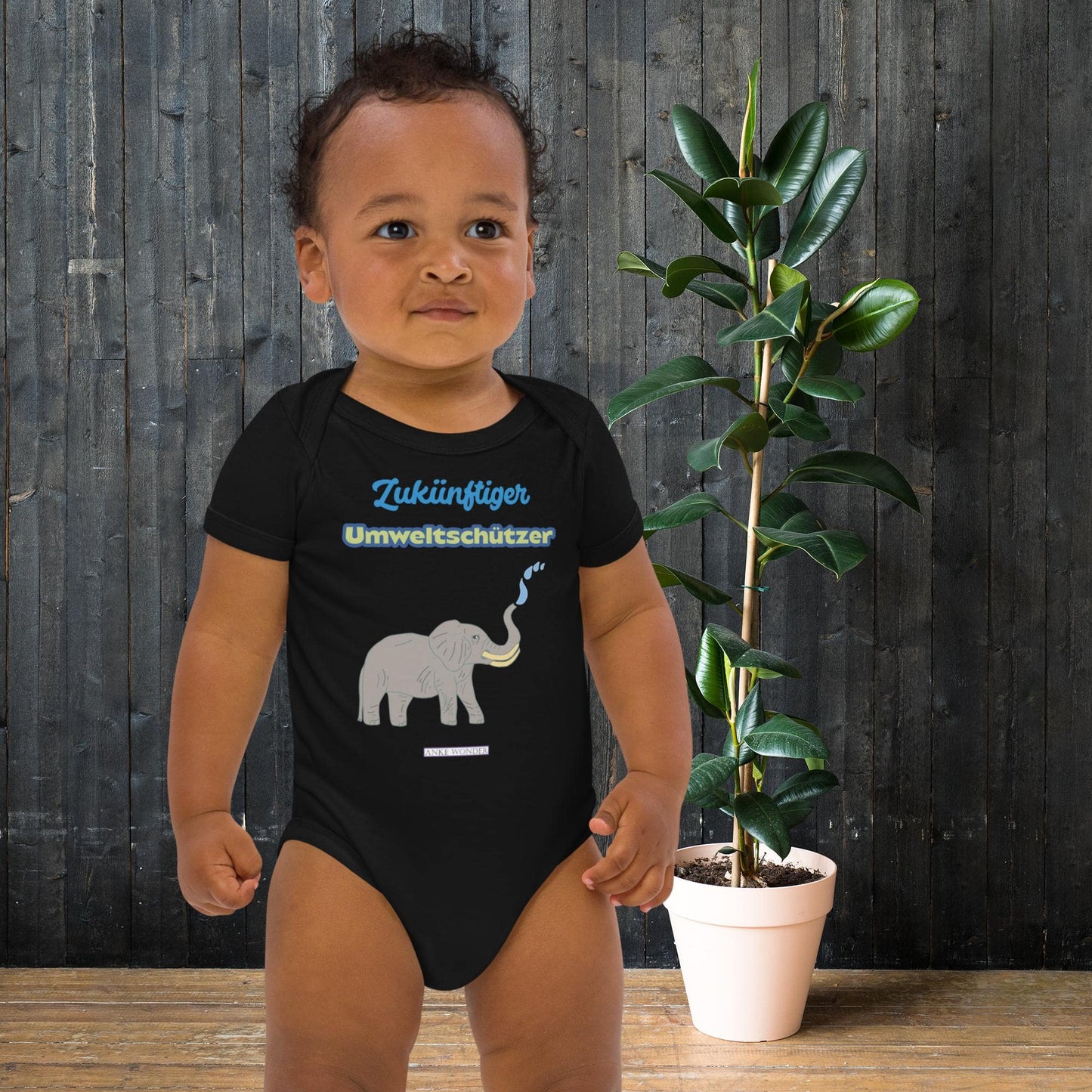 Child wearing a black onesie with elephant graphic and eco slogan 'Zukünftiger Umweltschützer', standing next to a potted plant against a wooden wall.