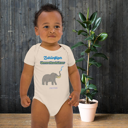 Baby boy wearing a onesie with elephant design and eco slogan 'Zukünftiger Umweltschützer' in front of a wooden wall and plant.