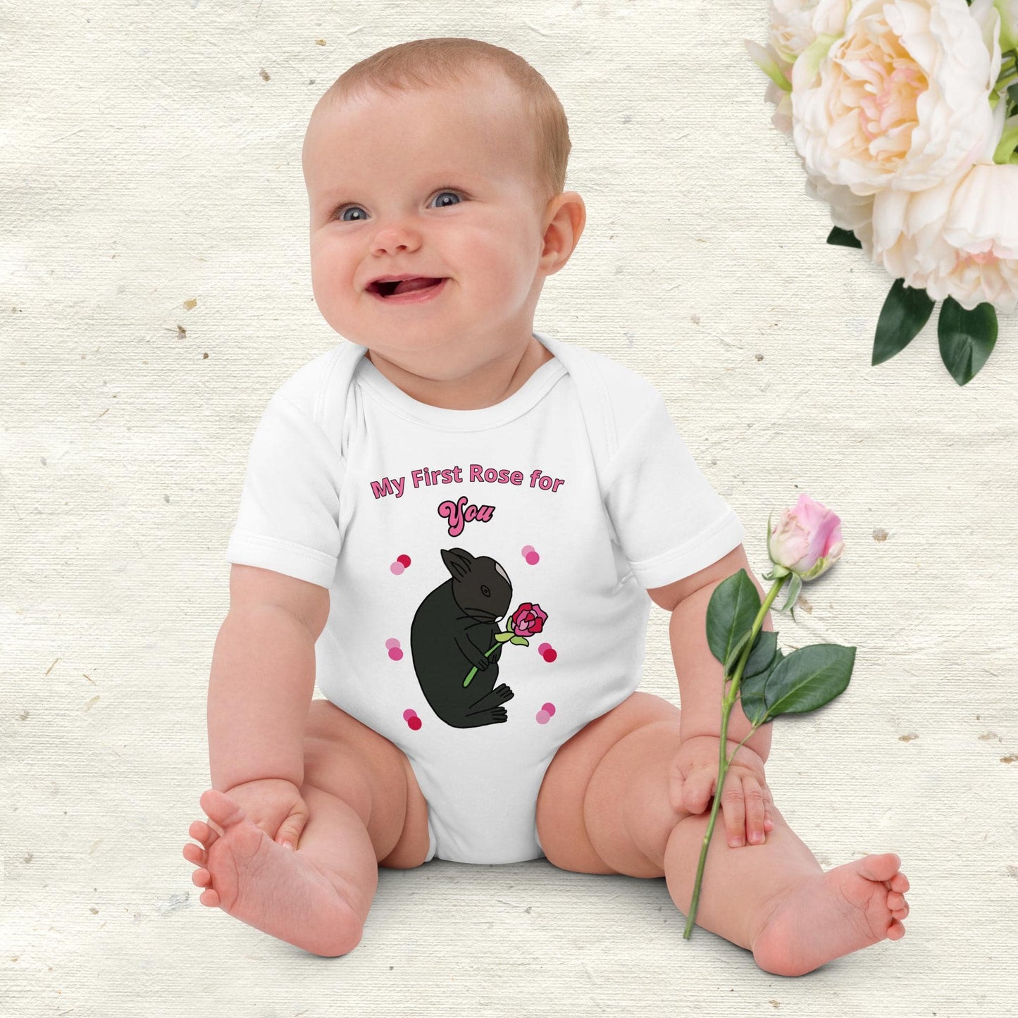Smiling baby girl sitting in a white bodysuit with bunny with rose design and decorative pink dots around the “My First Rose for You” print.