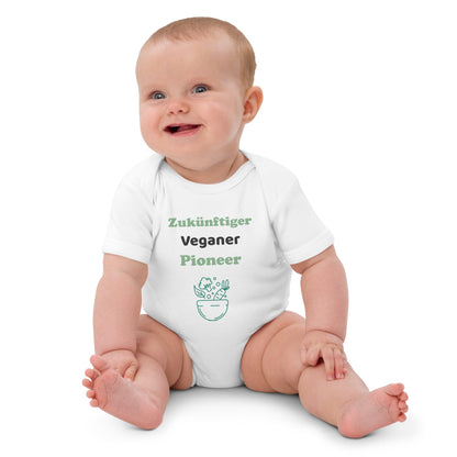 A baby girl sitting and wearing a white organic cotton bodysuit with "Zukünftiger Veganer Pionier" printed in green and black, smiling playfully.