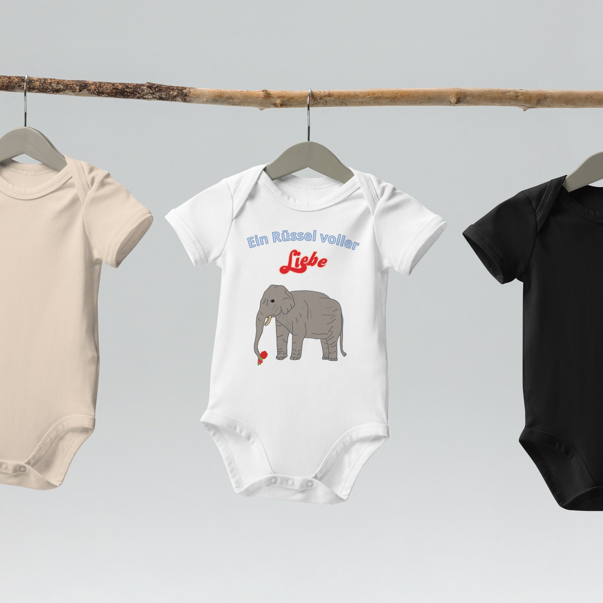  “Ein Rüssel voller Liebe” baby bodysuits with elephant design in white hanging on wooden hangers.