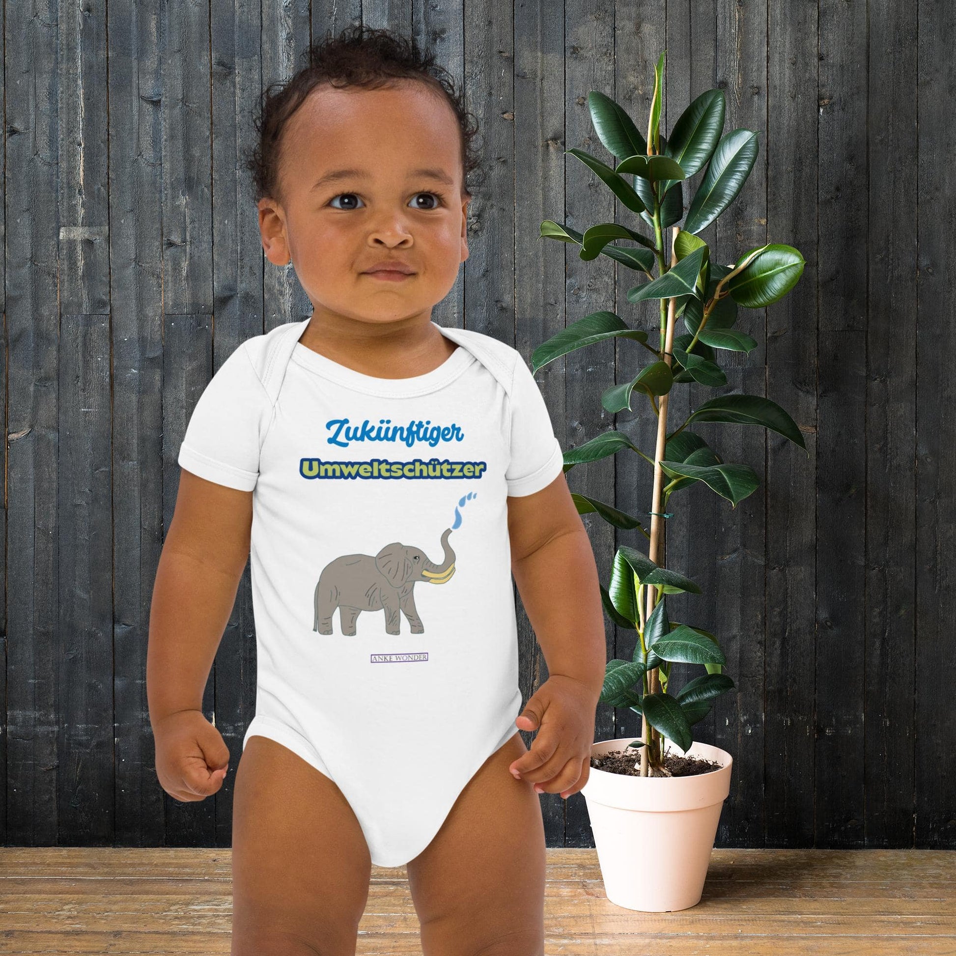 Baby boy wearing a white onesie with elephant design and eco slogan 'Zukünftiger Umweltschützer' in front of a wooden wall and plant.