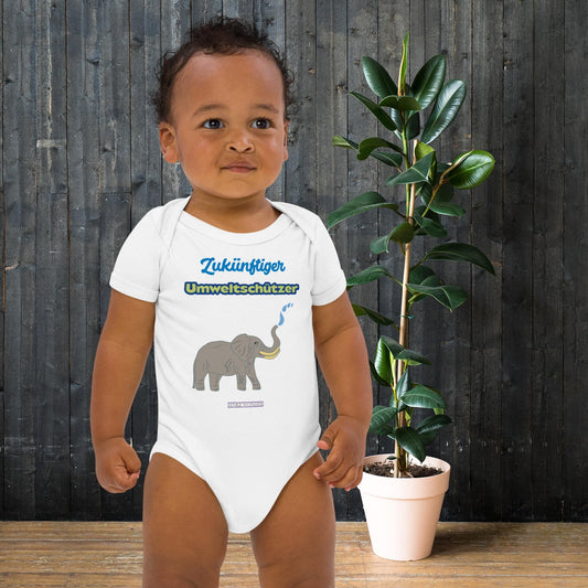 Baby boy wearing a white onesie with elephant design and eco slogan 'Zukünftiger Umweltschützer' in front of a wooden wall and plant.