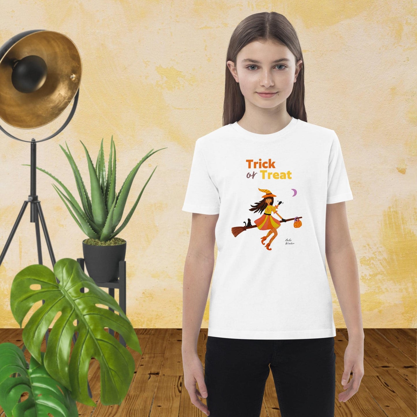 Girl wearing a 'Trick or Treat' t-shirt with a witch character in a room with plants and a lamp.