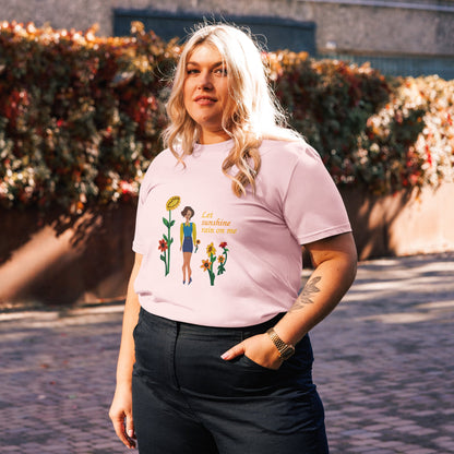 Woman wearing a pink t-shirt with a graphic design and'Let sunshine rain on me', standing outdoors.