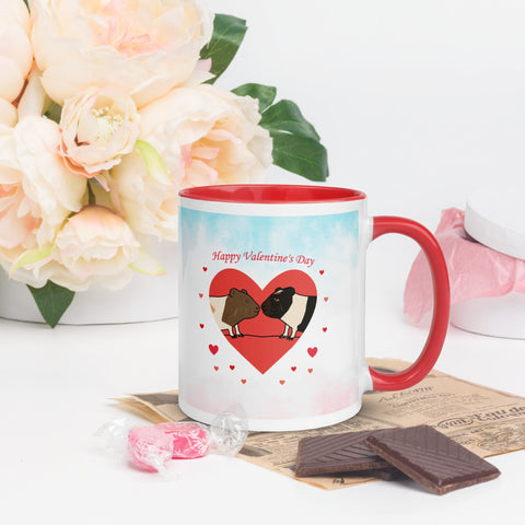 A Valentine's Day-themed ceramic mug with a red handle and inner rim, featuring an illustration of two guinea pigs inside a heart with small red hearts around them. The mug is placed on a table with chocolates, wrapped candies, and a bouquet of soft pink flowers in the background.