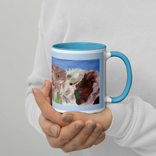 A person in a white long-sleeve top holds a ceramic mug with a blue interior and handle. The mug features a printed illustration of three guinea pigs in brown, white, and dark brown tones against a blue background.