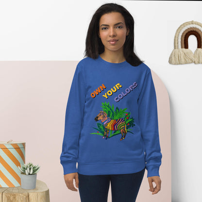 Woman wearing a blue sweatshirt with a colorful zebra design and text 'Own your colors", standing against a light pink wall.