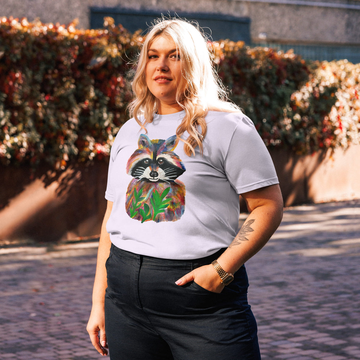 A woman wearing a white graphic t-shirt with a colorful raccoon print, paired with black pants, standing outdoors in front of a hedge.