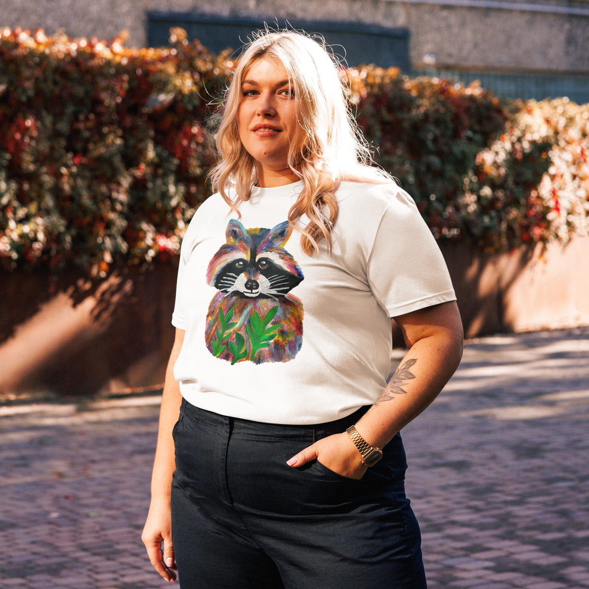 A woman wearing a white graphic t-shirt with a colorful raccoon print, paired with black pants, standing outdoors in front of a hedge.