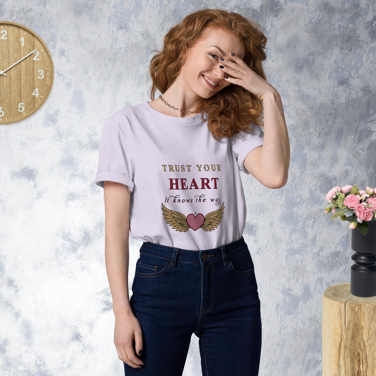 Woman wearing a lavender t-shirt with 'Trust Your Heart' text and wings graphic, standing against a gray wall with a clock and flowers.