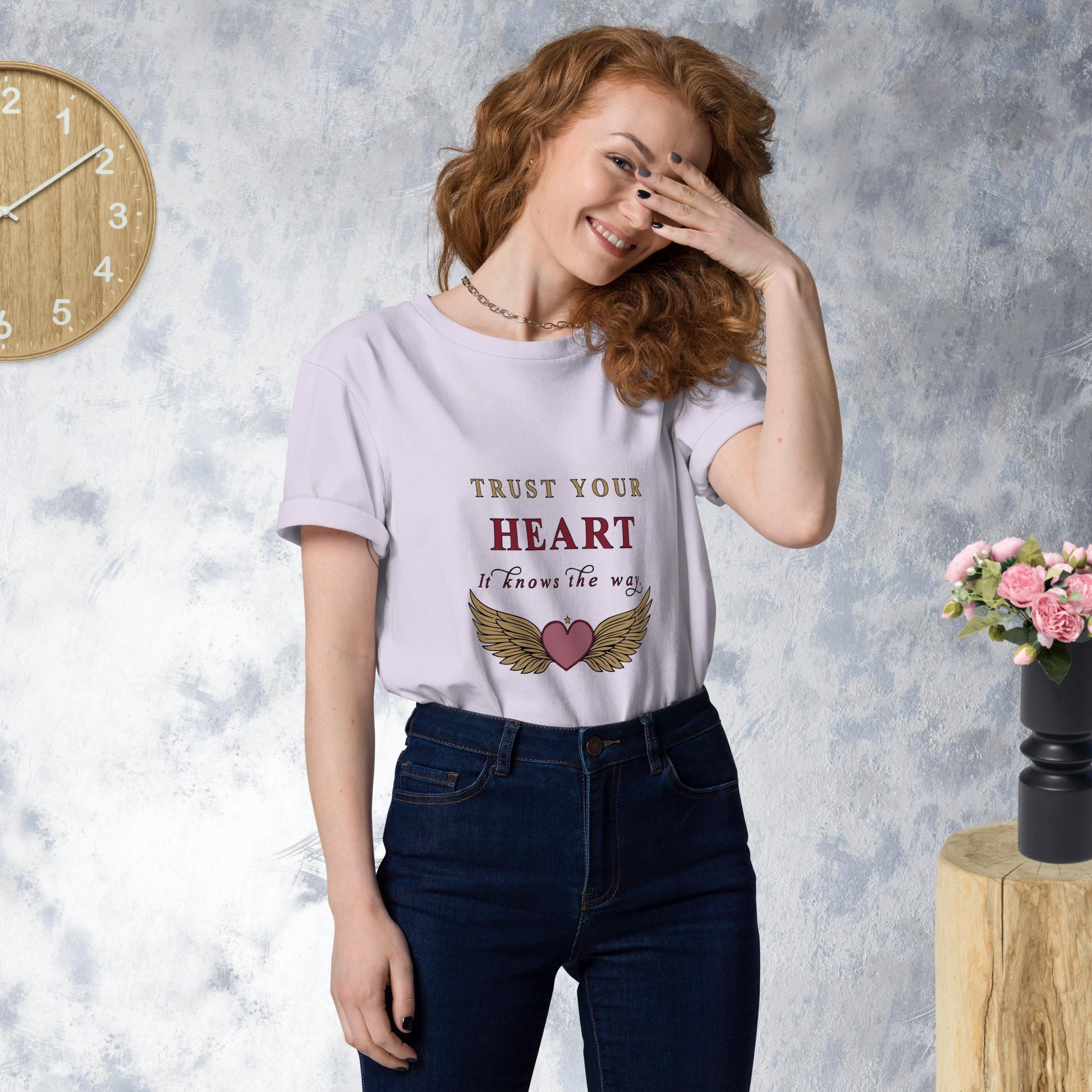 Woman wearing a lavender t-shirt with 'Trust Your Heart' text and wings graphic, standing against a gray wall with a clock and flowers.