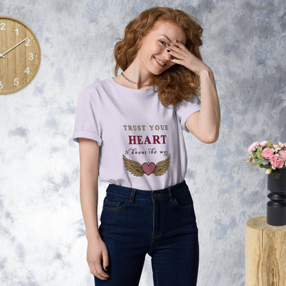 Woman wearing a lavender t-shirt with 'Trust Your Heart' text and wings graphic, standing against a gray wall with a clock and flowers.