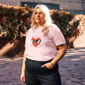 A plus-size woman with blonde wavy hair wearing a pink organic cotton Valentine's T-shirt featuring a cute guinea pig heart print, posing outdoors with one hand in her pocket and a warm smile.