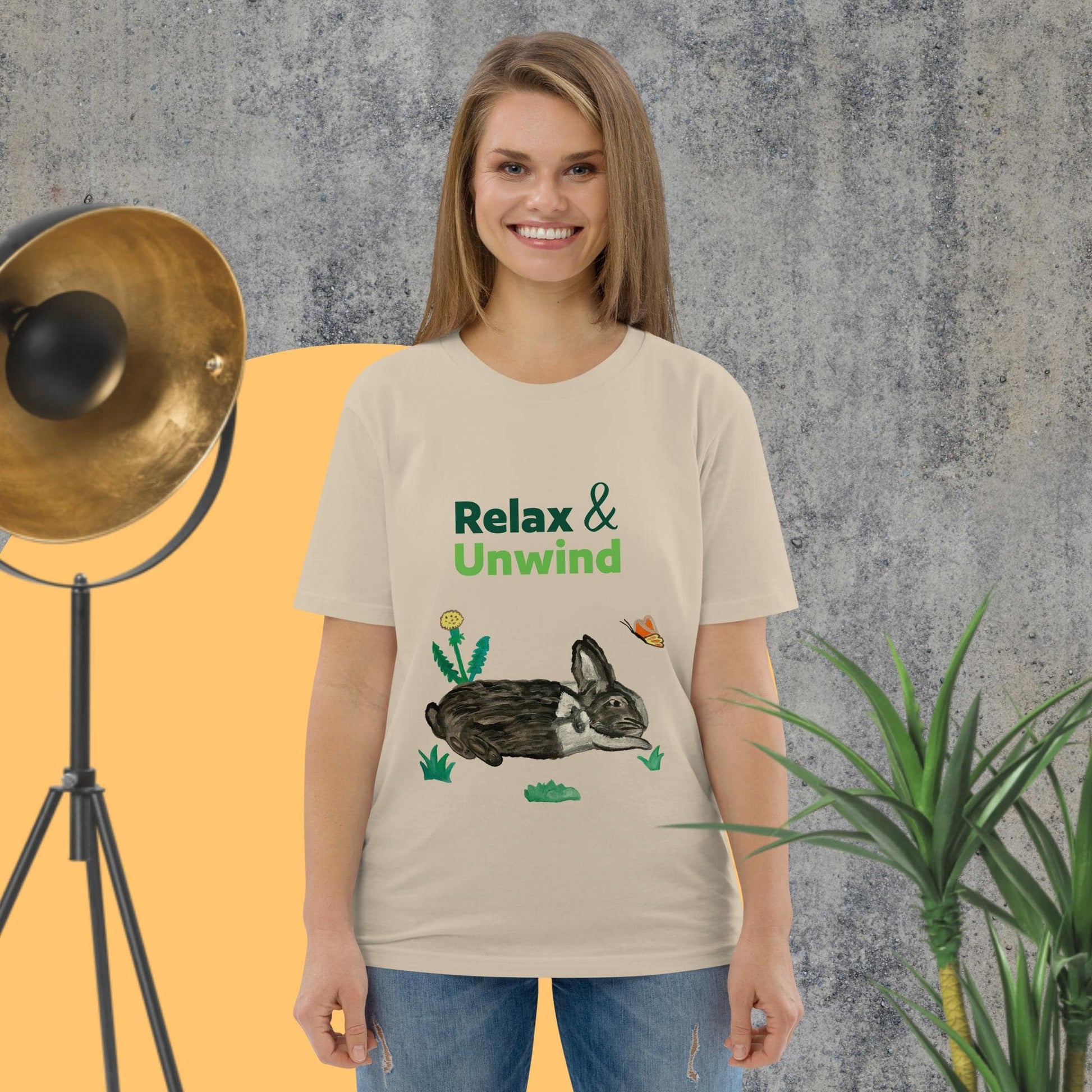 Woman wearing desert dust organic cotton t-shirt with hand-painted black-and-white rabbit, butterfly, greens, and "Relax & Unwind" text — front view.