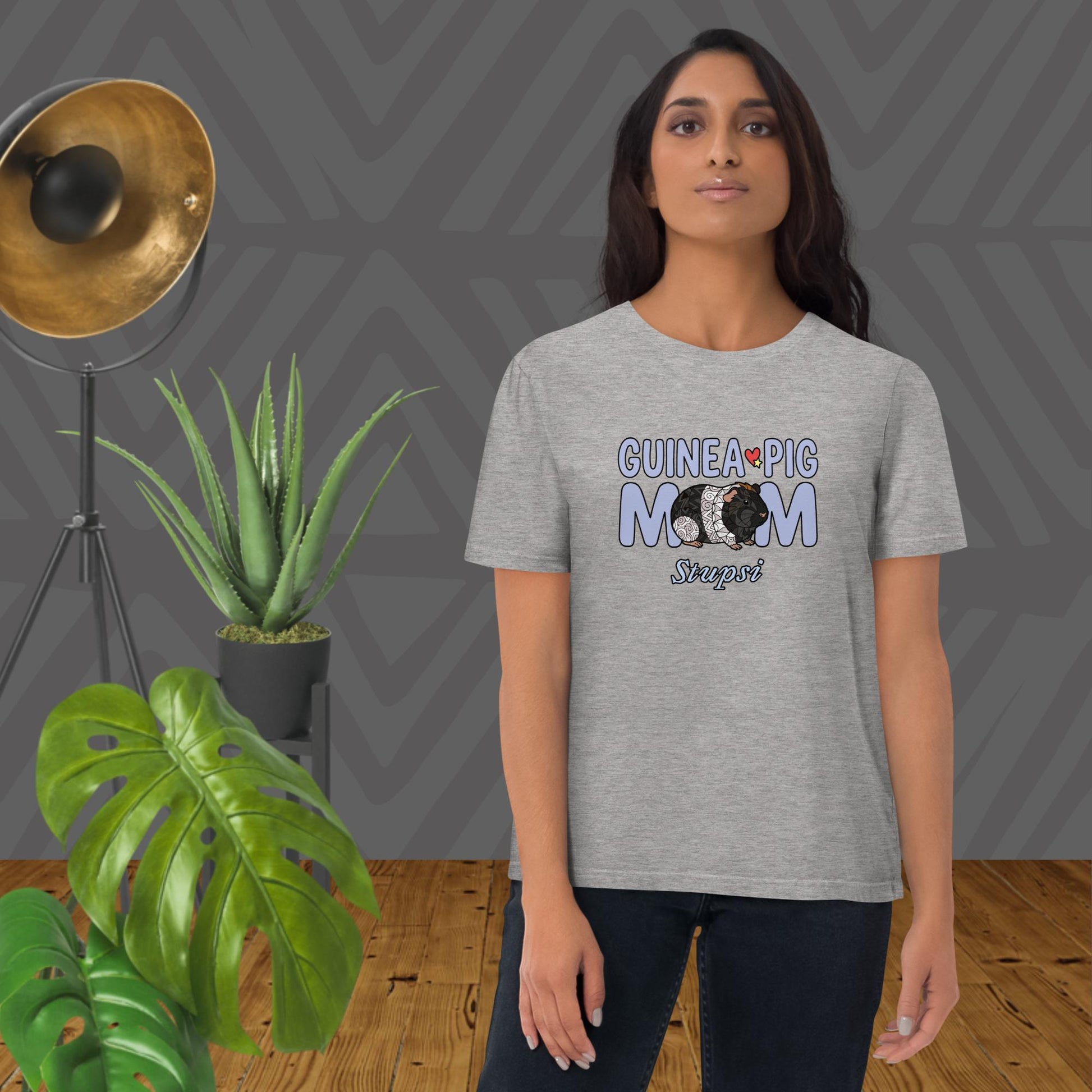 Woman wearing a gray t-shirt with 'Guinea Pig Mom' text and graphic, standing in a room with plants and a lamp.