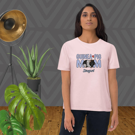 Woman wearing a pink t-shirt with 'Guinea Pig Mom' design, standing in a room with plants and a lamp.