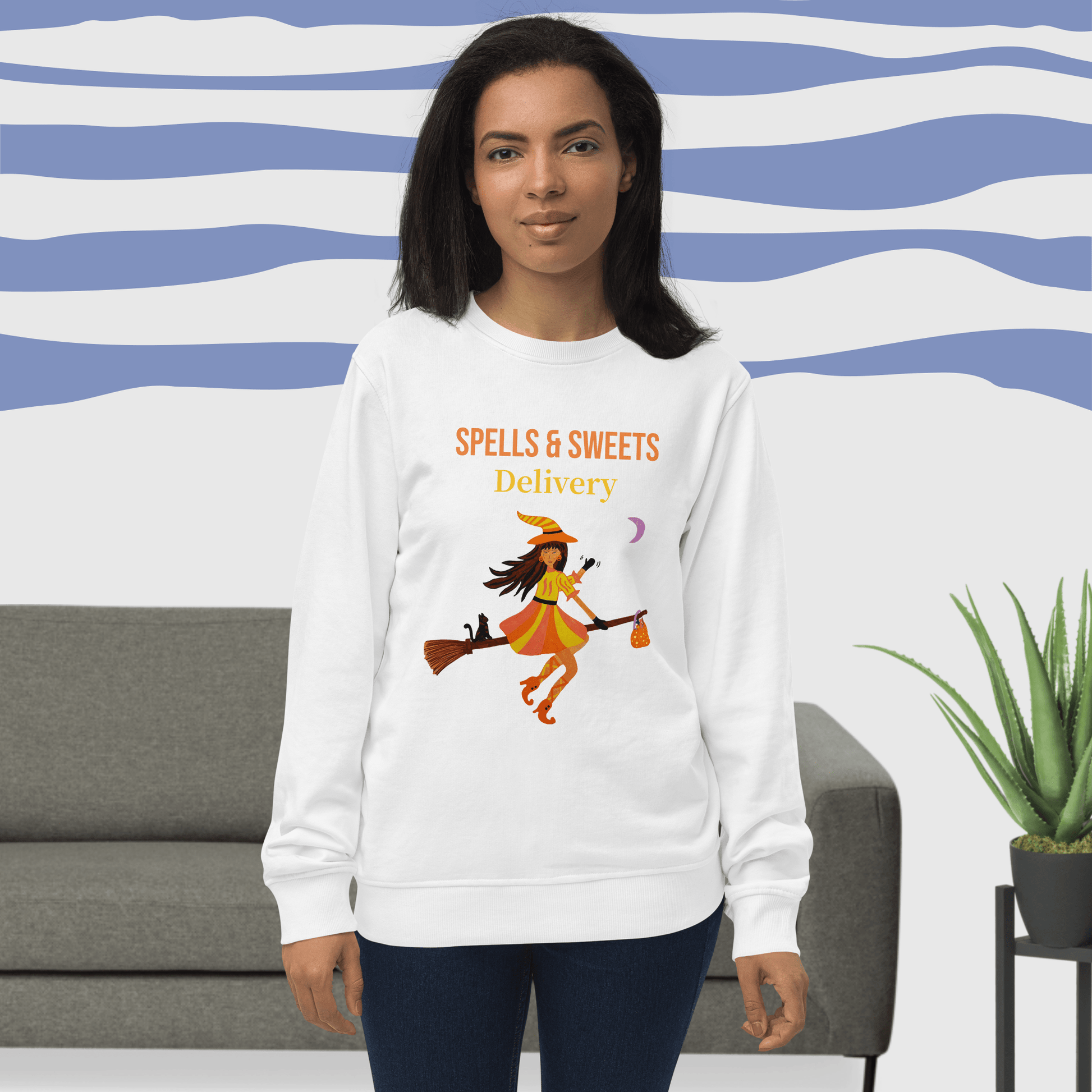 Woman wearing a white sweatshirt with 'Spells & Sweets Delivery' Halloween design in a living room setting.