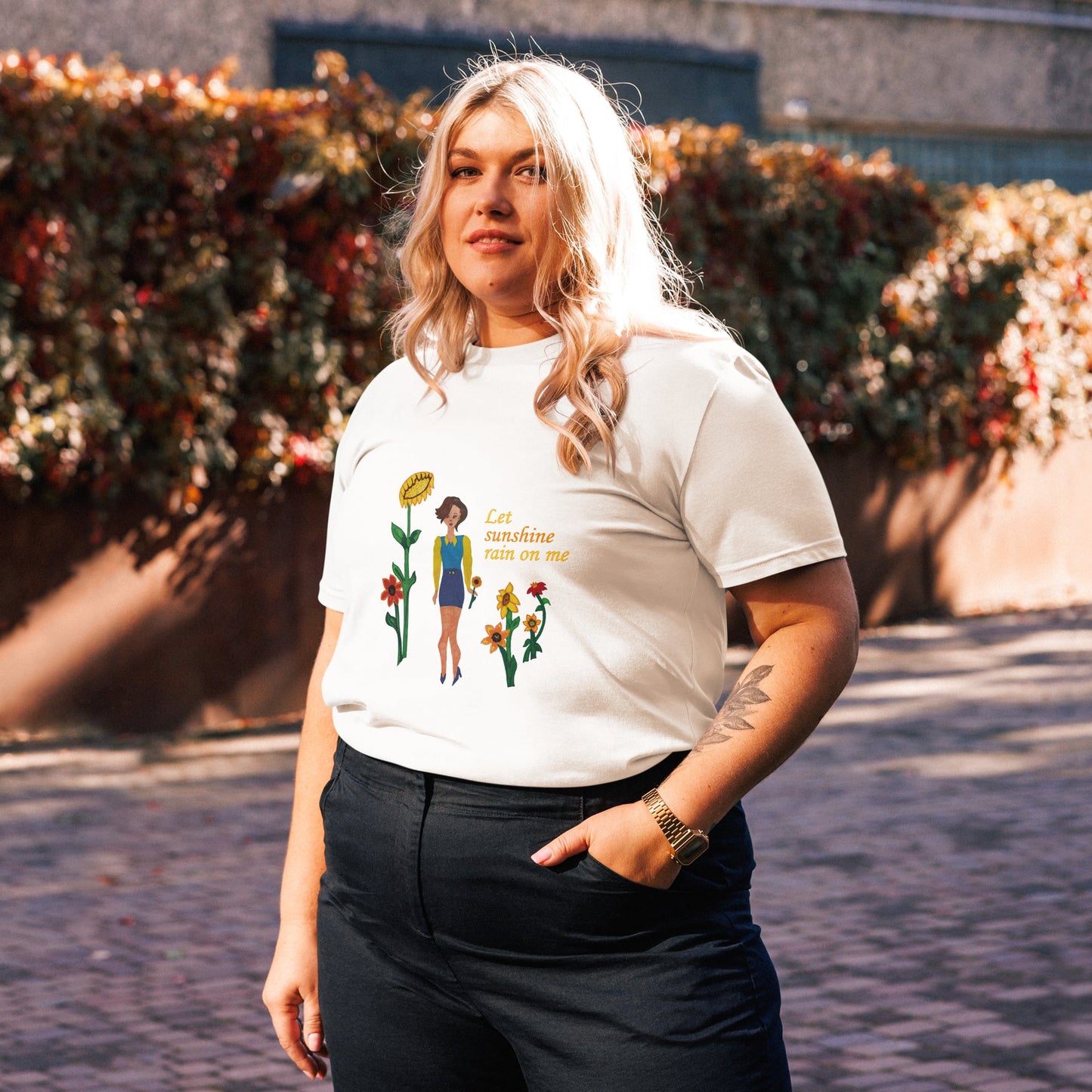Woman wearing a white t-shirt with a graphic design and text 'Let sunshine rain on me', standing outdoors.