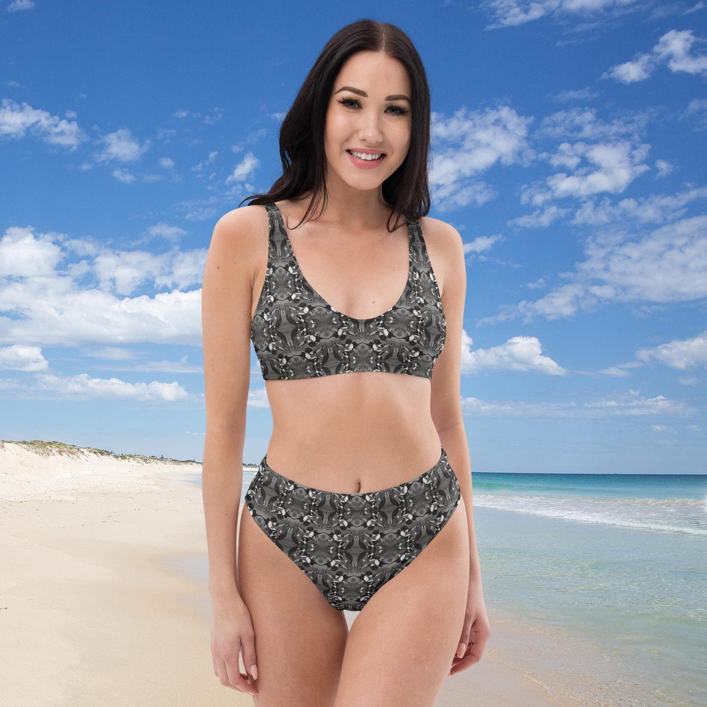 Woman in a gray bird patterned bikini standing on a beach with blue sky and ocean in the background.