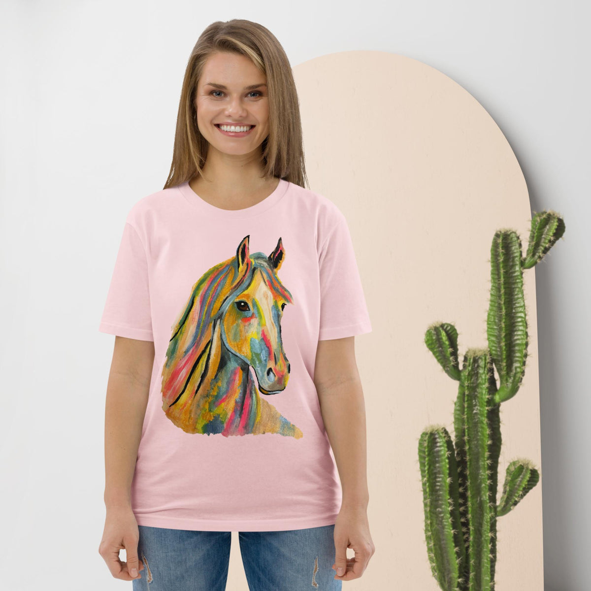A woman wearing a light pink t-shirt with a colorful watercolor-style horse graphic on the front, paired with blue jeans, standing in front of a beige arched backdrop with a cactus prop.