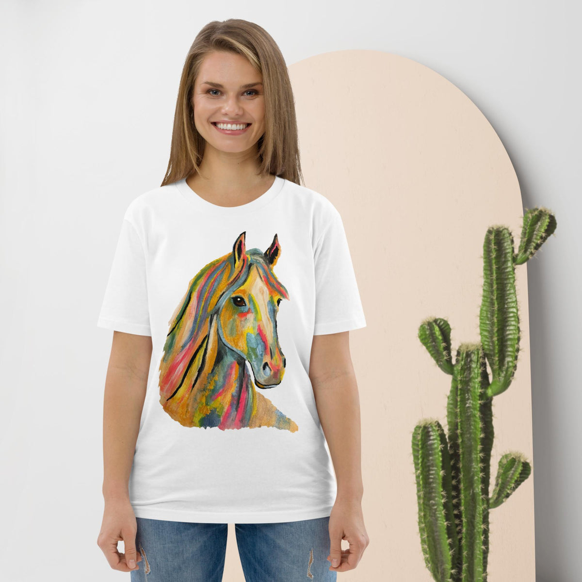 A woman wearing a white short-sleeve t-shirt with a colorful watercolor-style horse head graphic on the front, paired with blue jeans, standing against a light gray wall with a beige arch backdrop and a cactus prop.