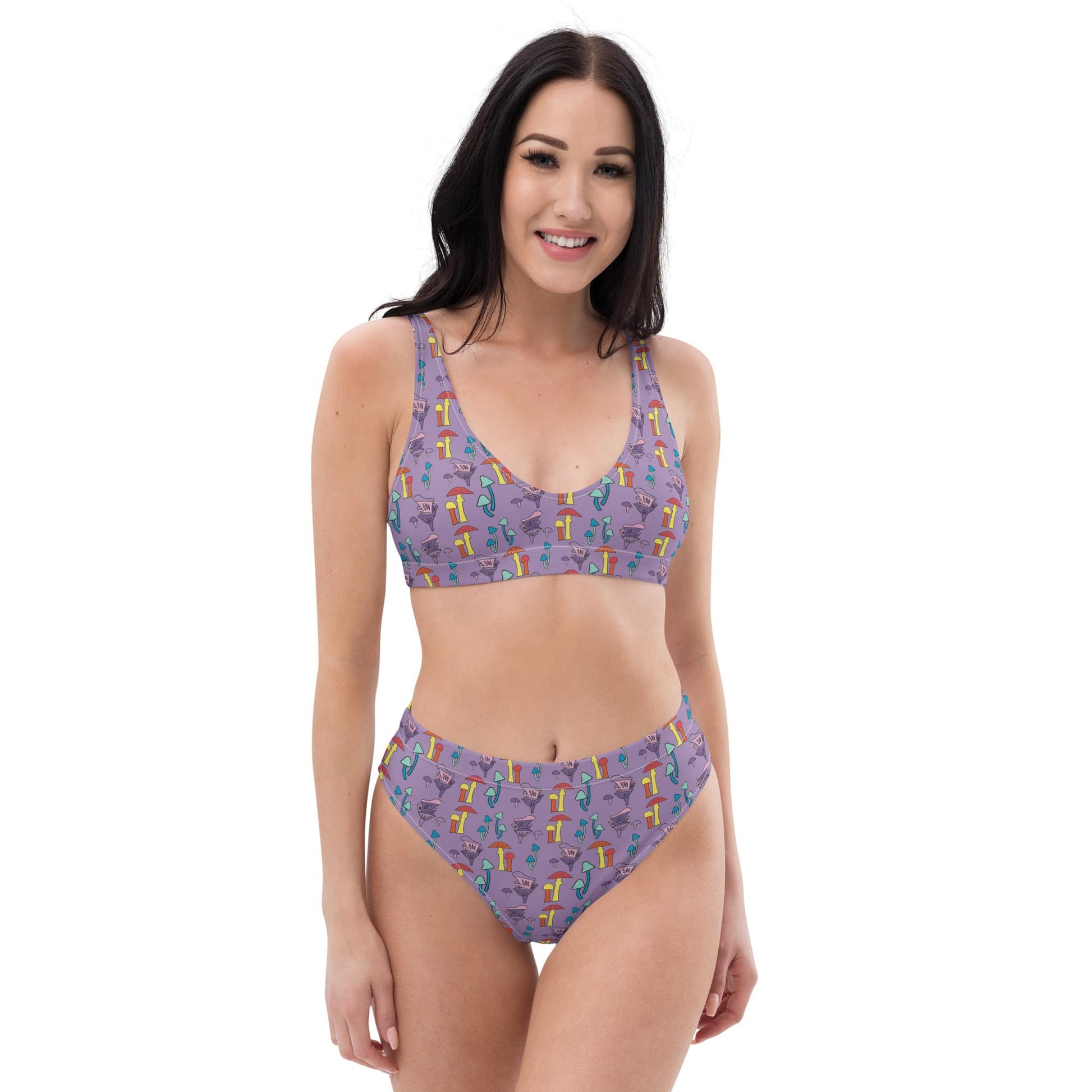 A woman models a high-waisted bikini with a whimsical, hand-painted purple mushroom print, made from recycled fabric, offering a flattering fit.