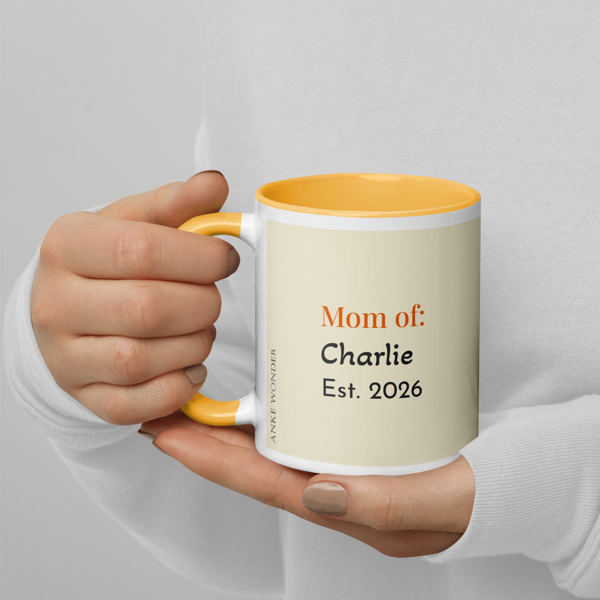 Hand holding a mug with text 'Mom of: Charlie Est. 2026' against a light gray background